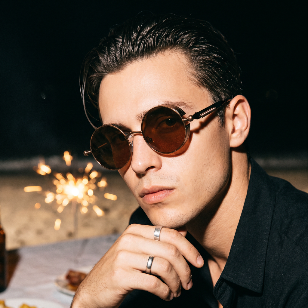 Man wearing round steampunk sunglasses at a sparkler-lit beach table at midnight