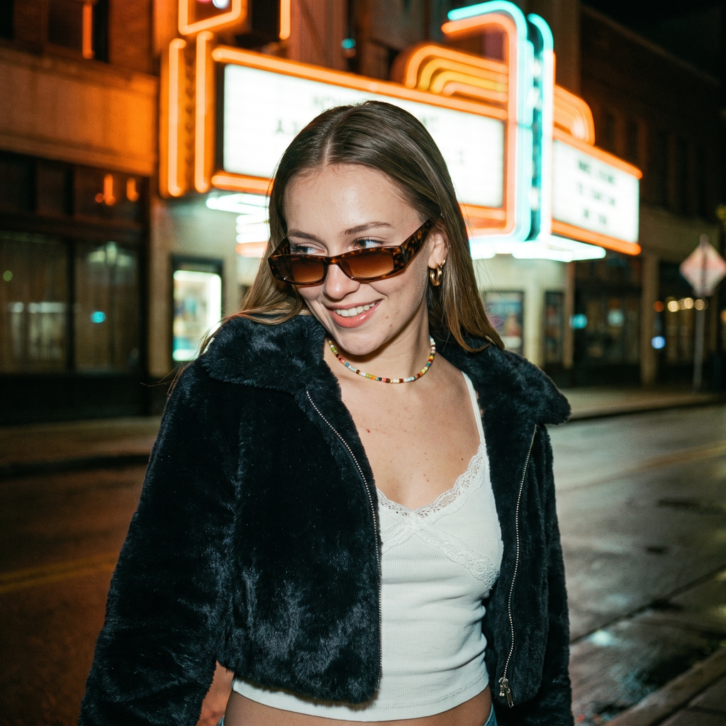 Woman on neon street in faux-fur wearing slim leopard rectangle Y2K sunglasses with brown lens
